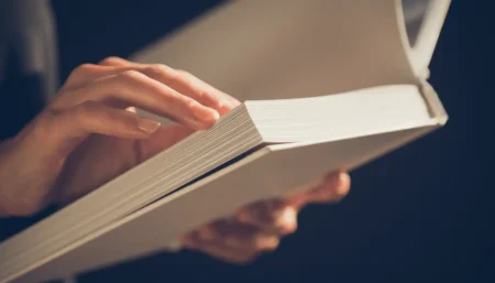 Detailný záber na ruky, ktoré držia a jemne otvárajú čerstvo vytlačenú knihu v mäkkej väzbe