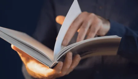 Detailný záber na mužské ruky, ktoré držia a jemne otvárajú čerstvo vytlačenú knihu v mäkkej väzbe
